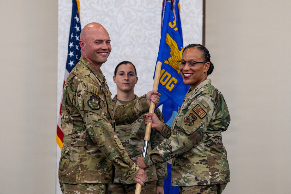 Two U.S. Air Force military personnel are participating in a formal change of command or assumption of command ceremony, jointly holding a blue guidon/flag. The flag displays 'MDG' (Medical Group) and a gold eagle emblem, indicating this is a medical group unit ceremony. A third service member stands at attention in the background.