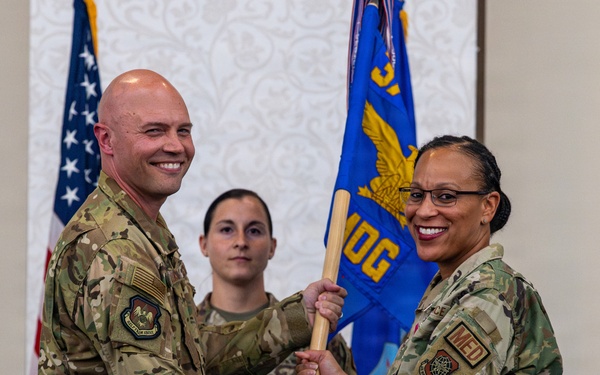 Two U.S. Air Force military personnel are participating in a formal change of command or assumption of command ceremony, jointly holding a blue guidon/flag. The flag displays 'MDG' (Medical Group) and a gold eagle emblem, indicating this is a medical group unit ceremony. A third service member stands at attention in the background.