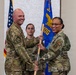 Two U.S. Air Force military personnel are participating in a formal change of command or assumption of command ceremony, jointly holding a blue guidon/flag. The flag displays 'MDG' (Medical Group) and a gold eagle emblem, indicating this is a medical group unit ceremony. A third service member stands at attention in the background.