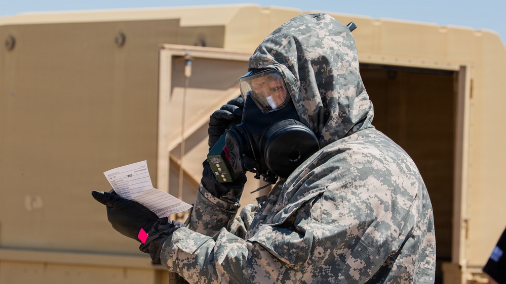 A military personnel wearing full CBRN (Chemical, Biological, Radiological, and Nuclear) protective gear, including a gas mask and digital camouflage uniform, is reviewing a document while holding a detection device. The individual is positioned outdoors near a tan/beige military structure or vehicle. The scene appears to be a training exercise or operational readiness drill.