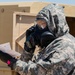 A military personnel wearing full CBRN (Chemical, Biological, Radiological, and Nuclear) protective gear, including a gas mask and digital camouflage uniform, is reviewing a document while holding a detection device. The individual is positioned outdoors near a tan/beige military structure or vehicle. The scene appears to be a training exercise or operational readiness drill.
