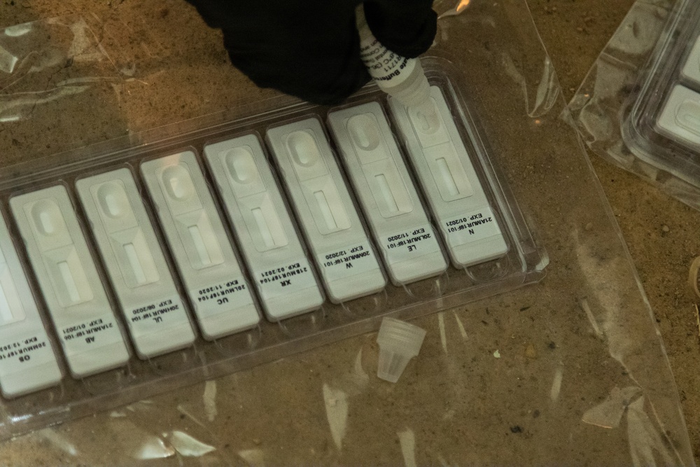The image shows a tray of rapid diagnostic test (RDT) cassettes arranged in a clear plastic holder, with a gloved hand holding a dropper or reagent bottle above one of the cassettes. Each cassette appears to be labeled with lot numbers and expiration dates ranging from 2020 to 2021. The setting appears to be a field or clinical testing environment based on the sandy/dusty surface visible in the background.