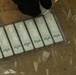 The image shows a tray of rapid diagnostic test (RDT) cassettes arranged in a clear plastic holder, with a gloved hand holding a dropper or reagent bottle above one of the cassettes. Each cassette appears to be labeled with lot numbers and expiration dates ranging from 2020 to 2021. The setting appears to be a field or clinical testing environment based on the sandy/dusty surface visible in the background.