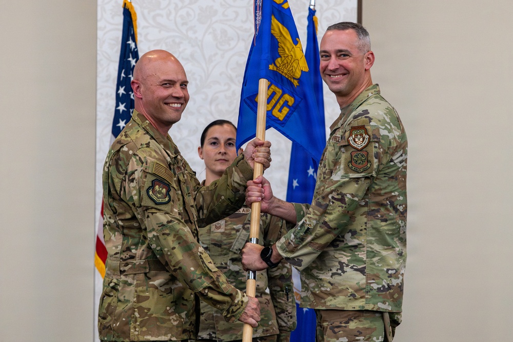 Two U.S. Air Force personnel in OCP (Operational Camouflage Pattern) uniforms are conducting a guidon/pennant passing ceremony, likely a change of command. A blue unit guidon bearing an eagle emblem and partial text 'OG' is being transferred between the two individuals. A third service member stands in the background observing the ceremony.