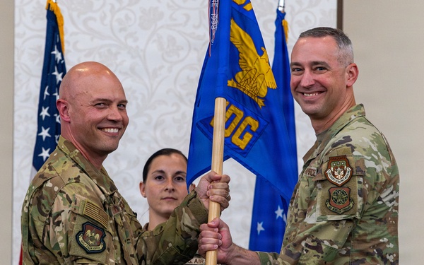 Two U.S. Air Force personnel in OCP (Operational Camouflage Pattern) uniforms are conducting a guidon/pennant passing ceremony, likely a change of command. A blue unit guidon bearing an eagle emblem and partial text 'OG' is being transferred between the two individuals. A third service member stands in the background observing the ceremony.