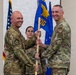 Two U.S. Air Force personnel in OCP (Operational Camouflage Pattern) uniforms are conducting a guidon/pennant passing ceremony, likely a change of command. A blue unit guidon bearing an eagle emblem and partial text 'OG' is being transferred between the two individuals. A third service member stands in the background observing the ceremony.