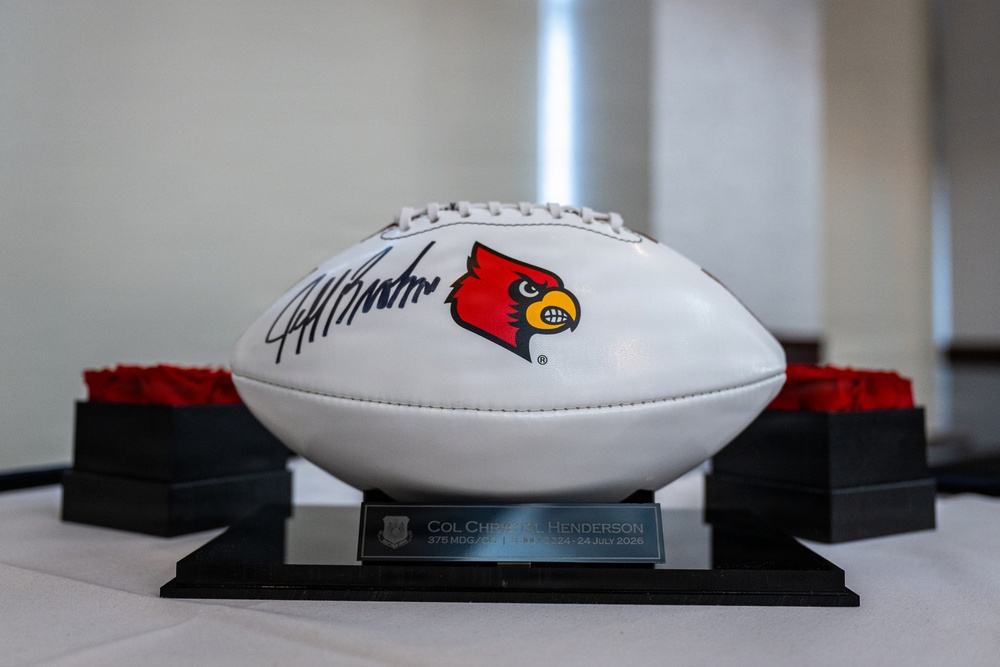 A white commemorative football featuring the University of Louisville Cardinals logo and an autograph is displayed on a black trophy stand. The plaque on the base reads 'COL Christopher Henderson, 375 MDG/CC' with dates referencing July 2026. The football appears to be a presentation gift or award, displayed on a white tablecloth with decorative red floral arrangements in the background.