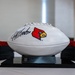 A white commemorative football featuring the University of Louisville Cardinals logo and an autograph is displayed on a black trophy stand. The plaque on the base reads 'COL Christopher Henderson, 375 MDG/CC' with dates referencing July 2026. The football appears to be a presentation gift or award, displayed on a white tablecloth with decorative red floral arrangements in the background.