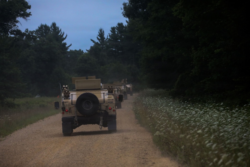 A convoy of military HMMWV (Humvee) vehicles is traveling along a narrow gravel road flanked by dense forest and vegetation. The image is taken from a rear perspective at dusk or dawn, capturing the vehicles moving away from the camera in a single-file formation. Dust is visible rising from the road surface behind the lead vehicle.