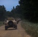 A convoy of military HMMWV (Humvee) vehicles is traveling along a narrow gravel road flanked by dense forest and vegetation. The image is taken from a rear perspective at dusk or dawn, capturing the vehicles moving away from the camera in a single-file formation. Dust is visible rising from the road surface behind the lead vehicle.