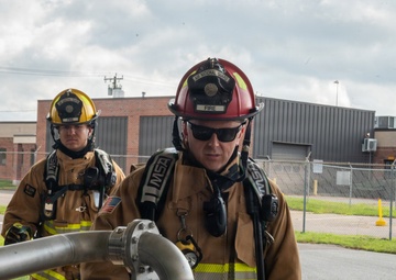 Three firefighters in full turnout gear are engaged in what appears to be a training exercise at a military or government facility. The foreground firefighter, wearing a red helmet marked 'FIRE' and an 'Air National Guard' insignia, is examining or operating a large metal pipe/hose assembly. The setting appears to be an outdoor area adjacent to industrial or military buildings.