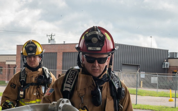 Three firefighters in full turnout gear are engaged in what appears to be a training exercise at a military or government facility. The foreground firefighter, wearing a red helmet marked 'FIRE' and an 'Air National Guard' insignia, is examining or operating a large metal pipe/hose assembly. The setting appears to be an outdoor area adjacent to industrial or military buildings.