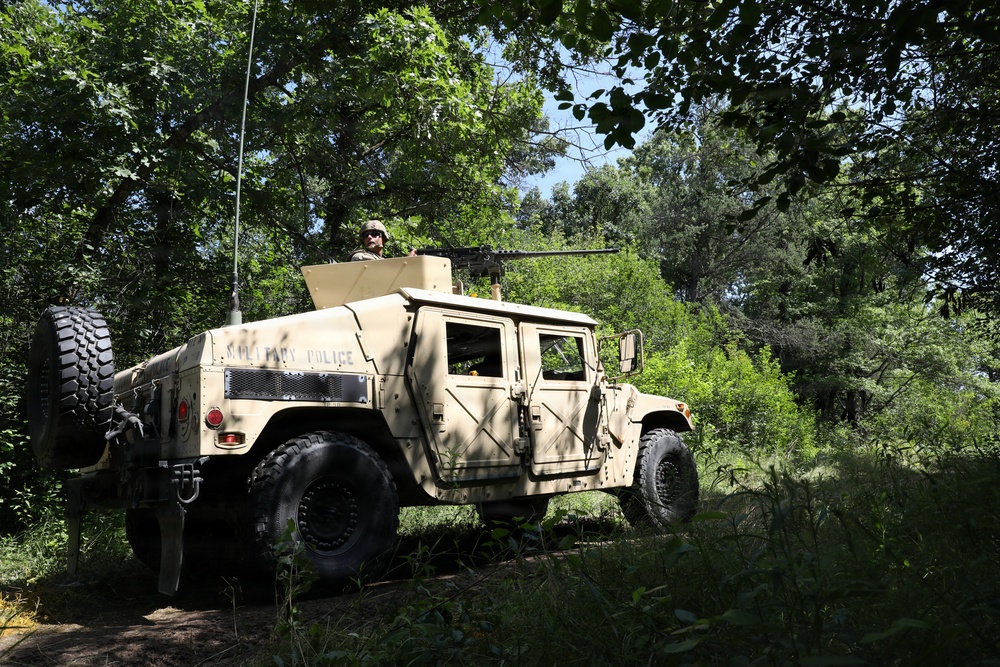 A tan/desert-colored Military Police HMMWV (Humvee) is positioned in a wooded area with dense green vegetation. A soldier is visible in the turret position atop the vehicle, with what appears to be a mounted weapon system. The vehicle bears 'MILITARY POLICE' markings on its rear quarter panel.