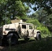 A tan/desert-colored Military Police HMMWV (Humvee) is positioned in a wooded area with dense green vegetation. A soldier is visible in the turret position atop the vehicle, with what appears to be a mounted weapon system. The vehicle bears 'MILITARY POLICE' markings on its rear quarter panel.