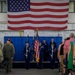 A formal military ceremony is taking place inside a large hangar or indoor facility, with a color guard presenting flags before an audience of military personnel and civilians. A large American flag is prominently displayed on the wall behind the stage. The setting and uniforms suggest a U.S. Air Force change of command or similar official ceremony.