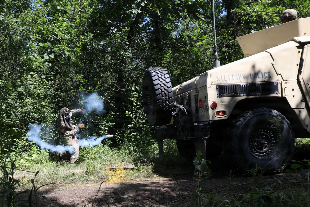 A military training exercise is taking place in a wooded area involving at least two personnel and a Military Police HMMWV (Humvee). One soldier appears to be deploying or throwing a smoke grenade, producing visible blue-gray smoke near the rear of the vehicle. A second individual is visible in the turret position atop the Humvee.