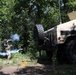 A military training exercise is taking place in a wooded area involving at least two personnel and a Military Police HMMWV (Humvee). One soldier appears to be deploying or throwing a smoke grenade, producing visible blue-gray smoke near the rear of the vehicle. A second individual is visible in the turret position atop the Humvee.