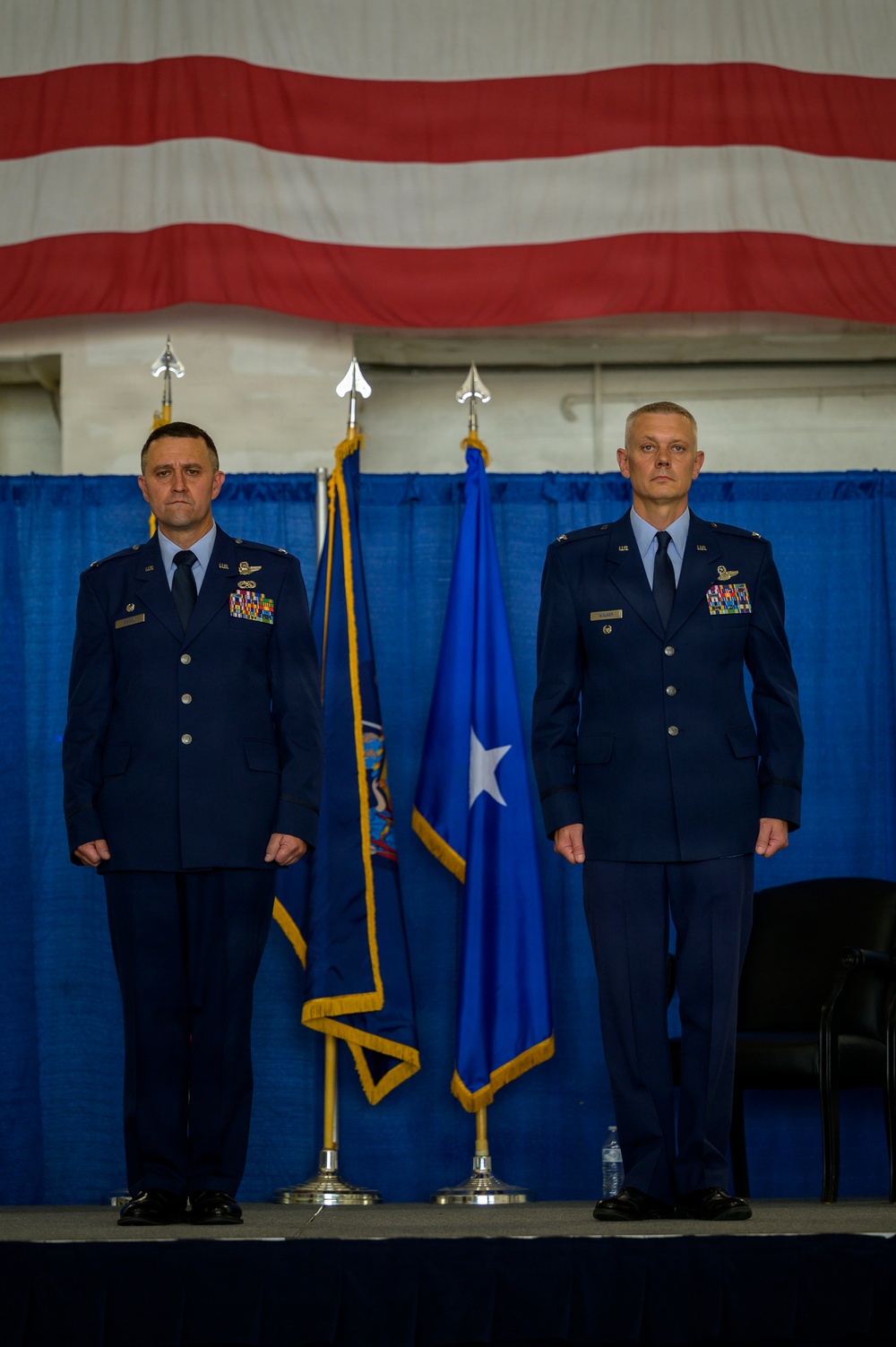 Two U.S. Air Force officers in dress blue uniforms stand at attention on a stage during a formal military ceremony. A large American flag is displayed as a backdrop along with blue ceremonial flags with gold fringe, one bearing a single white star indicating a one-star general rank. The setting appears to be inside a hangar or large indoor facility decorated for an official event such as a change of command ceremony.