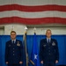 Two U.S. Air Force officers in dress blue uniforms stand at attention on a stage during a formal military ceremony. A large American flag is displayed as a backdrop along with blue ceremonial flags with gold fringe, one bearing a single white star indicating a one-star general rank. The setting appears to be inside a hangar or large indoor facility decorated for an official event such as a change of command ceremony.