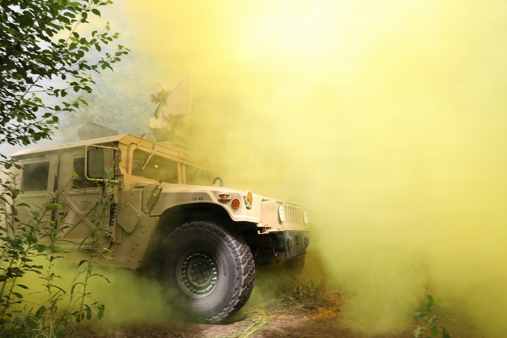 A military HMMWV (High Mobility Multipurpose Wheeled Vehicle), commonly known as a Humvee, is photographed on a dirt trail surrounded by dense yellow-green smoke, likely from a smoke grenade used in a training exercise. The vehicle appears to be tan/desert-colored and is equipped with a roof-mounted weapon system or turret. The scene suggests a military field training exercise conducted in a wooded or vegetated environment.