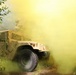 A military HMMWV (High Mobility Multipurpose Wheeled Vehicle), commonly known as a Humvee, is photographed on a dirt trail surrounded by dense yellow-green smoke, likely from a smoke grenade used in a training exercise. The vehicle appears to be tan/desert-colored and is equipped with a roof-mounted weapon system or turret. The scene suggests a military field training exercise conducted in a wooded or vegetated environment.
