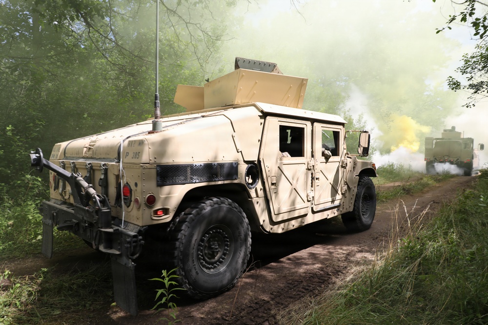 A tan/desert-camouflaged HMMWV (Humvee) military vehicle is traveling along a dirt trail through a wooded area. Yellow and white smoke is visible in the background, consistent with smoke grenades or screening smoke used in military training exercises. A second military vehicle is partially visible behind the smoke in the background.