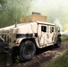 A tan/desert-camouflaged HMMWV (Humvee) military vehicle is traveling along a dirt trail through a wooded area. Yellow and white smoke is visible in the background, consistent with smoke grenades or screening smoke used in military training exercises. A second military vehicle is partially visible behind the smoke in the background.