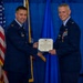 Two United States Air Force officers in dress blue uniforms are jointly holding an official certificate or award document during a formal military ceremony. The officer on the right is wearing a medal, suggesting he is the recipient of the award being presented. The event appears to take place in a decorated venue with American and Air Force flags visible in the background.