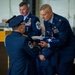 A formal U.S. Air Force military ceremony is taking place indoors, likely a retirement or change of command ceremony. A colonel (identified by silver eagle insignia) is receiving a folded American flag from a Master Sergeant wearing white ceremonial gloves. A Chief Master Sergeant stands in the background near a podium.
