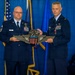 Two U.S. Air Force personnel in dress blue uniforms are holding a large ceremonial Command Pilot wings plaque during what appears to be a military ceremony. The individual on the left is a Senior Non-Commissioned Officer (name tag reads 'MEADE') and the officer on the right holds the rank of Lieutenant Colonel. A third uniformed individual is seated in the background observing the ceremony.