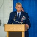 A U.S. Air Force officer in dress blues is speaking at a wooden podium equipped with a microphone. He is wearing multiple service ribbons, a medal, pilot wings, and rank insignia consistent with a senior officer. The setting appears to be an indoor military ceremony, likely a change of command or awards presentation.