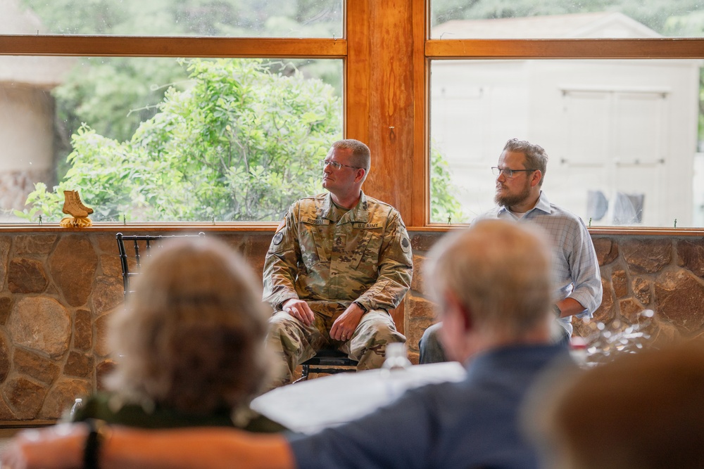 Two men are seated against a stone wall beneath large wooden-framed windows in what appears to be a meeting or presentation setting. One man is wearing a U.S. Army camouflage uniform (OCP pattern) with visible name tape, while the other is in civilian attire. An audience is visible in the foreground, suggesting a panel discussion or briefing is taking place.