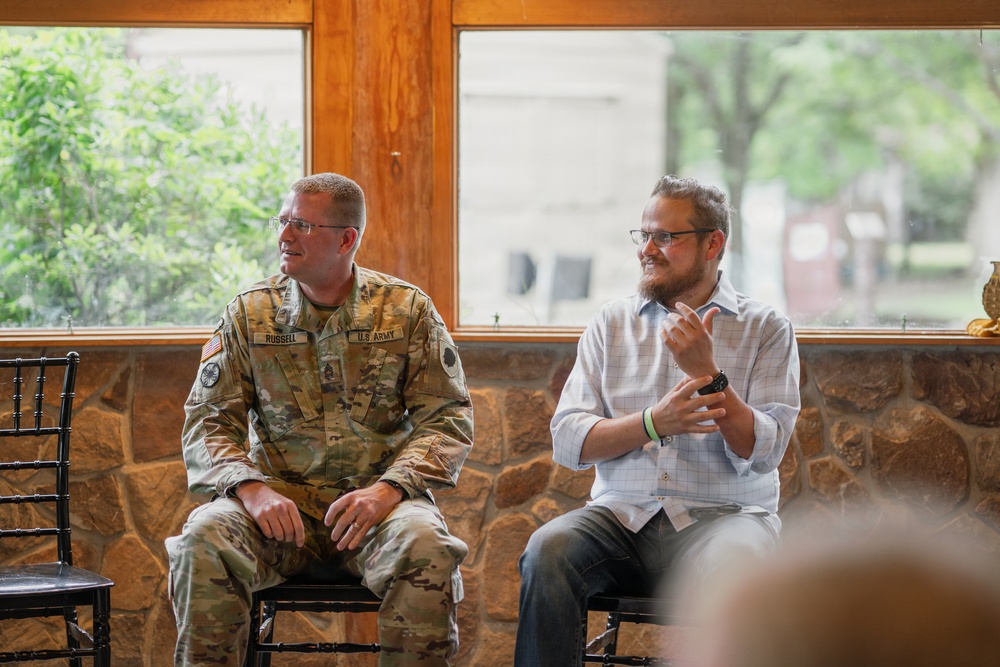 Two men are seated side by side in what appears to be an indoor venue with stone walls and large windows. One man is wearing a U.S. Army camouflage uniform with the name tape 'RUSSELL' visible, while the other is a bearded civilian in a light blue button-up shirt. Both appear to be participating in a panel discussion or presentation, with the civilian gesturing as if speaking.