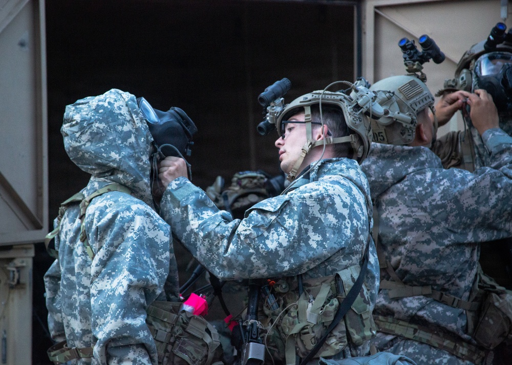 The image shows multiple soldiers in US Army Universal Camouflage Pattern (UCP) uniforms conducting what appears to be a CBRN (Chemical, Biological, Radiological, Nuclear) drill or preparation exercise. A central soldier wearing a tactical helmet with a night vision device mount is assisting another soldier in donning a gas mask. Additional soldiers in the background are also equipping or adjusting protective gear.