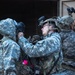 The image shows multiple soldiers in US Army Universal Camouflage Pattern (UCP) uniforms conducting what appears to be a CBRN (Chemical, Biological, Radiological, Nuclear) drill or preparation exercise. A central soldier wearing a tactical helmet with a night vision device mount is assisting another soldier in donning a gas mask. Additional soldiers in the background are also equipping or adjusting protective gear.