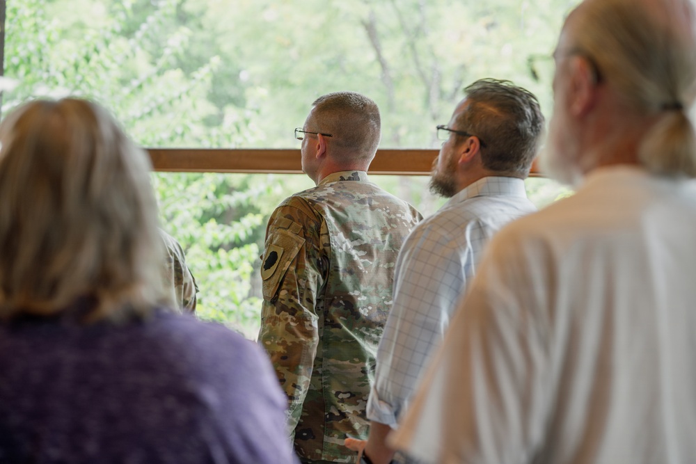 A group of four individuals are standing indoors near a large window overlooking a lush green outdoor area. One person is wearing a military camouflage uniform (OCP/MultiCam pattern), while the others are dressed in civilian clothing. They appear to be observing or attending a presentation or briefing together.