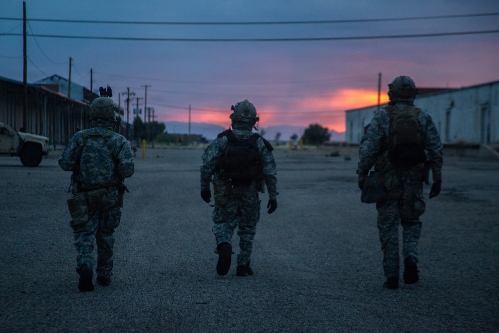 Three military personnel in full combat gear are walking away from the camera along a paved road at dusk or dawn. The scene appears to be at a military installation or training facility, with warehouse-style buildings on either side and a dramatic red-orange sunset on the horizon. The low-light conditions and silhouetted figures create a cinematic, atmospheric composition.