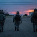 Three military personnel in full combat gear are walking away from the camera along a paved road at dusk or dawn. The scene appears to be at a military installation or training facility, with warehouse-style buildings on either side and a dramatic red-orange sunset on the horizon. The low-light conditions and silhouetted figures create a cinematic, atmospheric composition.