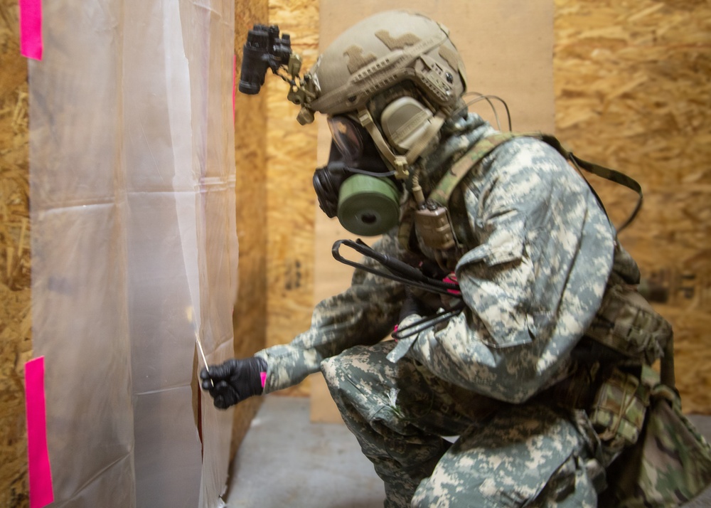 A military or law enforcement operator in full protective gear is crouching and cutting or breaching a plastic sheeting barrier secured with pink tape markers. The individual is equipped with a gas mask, tactical helmet with night vision mount, body armor, and digital camouflage uniform. The setting appears to be a training facility constructed with oriented strand board (OSB) walls.