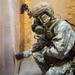 A military or law enforcement operator in full protective gear is crouching and cutting or breaching a plastic sheeting barrier secured with pink tape markers. The individual is equipped with a gas mask, tactical helmet with night vision mount, body armor, and digital camouflage uniform. The setting appears to be a training facility constructed with oriented strand board (OSB) walls.