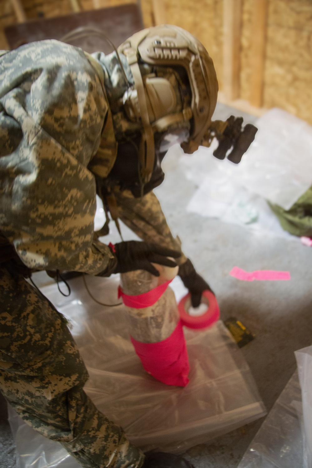 A soldier in digital camouflage uniform and tactical helmet is bent over, working with bright red/pink tape on what appears to be a cylindrical object placed on a plastic sheet. The setting appears to be an indoor training or operational environment with plywood walls visible in the background. The activity suggests a wrapping, sealing, or improvised explosive device (IED) training exercise.