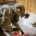 A soldier in digital camouflage uniform and tactical helmet is bent over, working with bright red/pink tape on what appears to be a cylindrical object placed on a plastic sheet. The setting appears to be an indoor training or operational environment with plywood walls visible in the background. The activity suggests a wrapping, sealing, or improvised explosive device (IED) training exercise.
