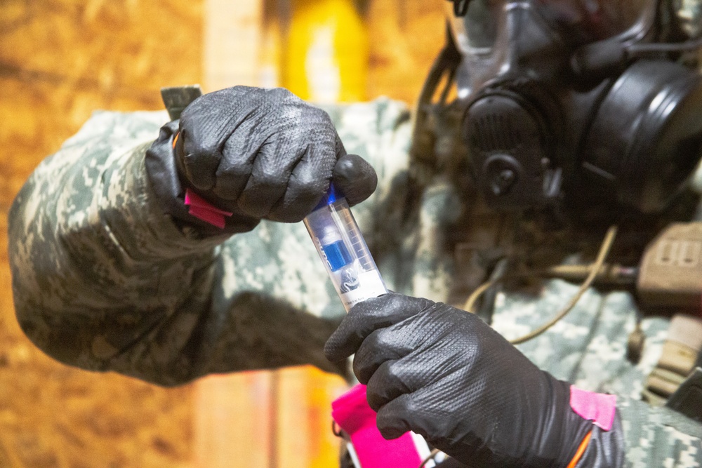 A person wearing military camouflage uniform, black protective gloves, and a full-face gas mask is handling a clear test tube or detection vial containing a blue substance. The activity appears to be a CBRN (Chemical, Biological, Radiological, Nuclear) detection or sampling procedure. Pink/magenta colored markers or tags are visible on the gloves and equipment, likely used for identification or procedural sequencing.
