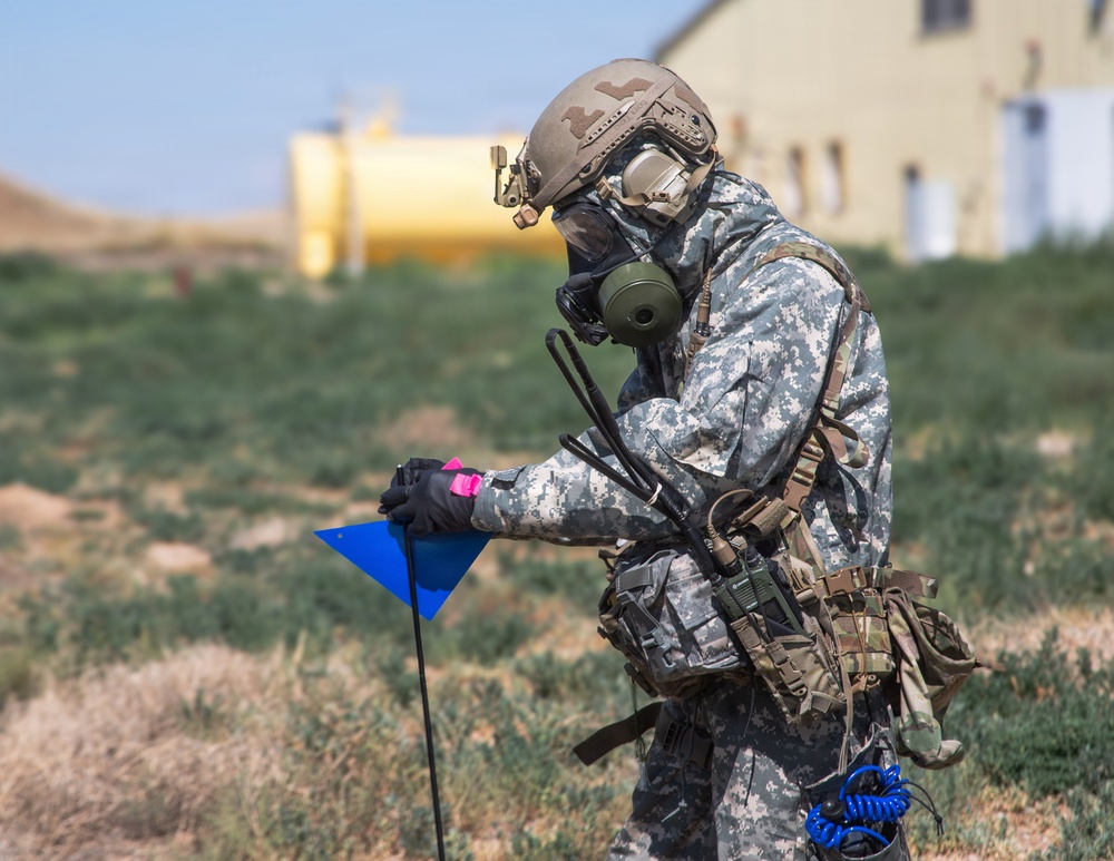 A military soldier in full CBRN (Chemical, Biological, Radiological, Nuclear) protective gear is conducting what appears to be a hazard marking or survey operation in an open field. The soldier is wearing a gas mask, tactical helmet, digital camouflage uniform, and carrying multiple communication devices while handling a blue triangular marker flag. The background shows a semi-arid training environment with structures and a yellow cylindrical tank visible.
