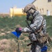 A military soldier in full CBRN (Chemical, Biological, Radiological, Nuclear) protective gear is conducting what appears to be a hazard marking or survey operation in an open field. The soldier is wearing a gas mask, tactical helmet, digital camouflage uniform, and carrying multiple communication devices while handling a blue triangular marker flag. The background shows a semi-arid training environment with structures and a yellow cylindrical tank visible.