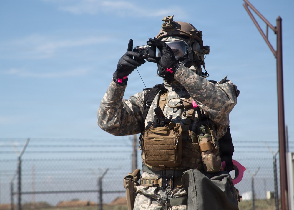 A military or law enforcement personnel in full CBRN (Chemical, Biological, Radiological, Nuclear) protective gear is photographing or recording with a handheld camera. The individual is wearing US Army ACU (Army Combat Uniform) digital camouflage, a full-face gas mask, tactical helmet, and MOLLE-equipped chest rig. The setting appears to be an outdoor training or operational environment near a chain-link fence with barbed wire.