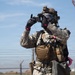 A military or law enforcement personnel in full CBRN (Chemical, Biological, Radiological, Nuclear) protective gear is photographing or recording with a handheld camera. The individual is wearing US Army ACU (Army Combat Uniform) digital camouflage, a full-face gas mask, tactical helmet, and MOLLE-equipped chest rig. The setting appears to be an outdoor training or operational environment near a chain-link fence with barbed wire.