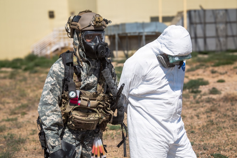 Two individuals are shown in full protective gear in an outdoor setting near a building. One person wears military digital camouflage uniform with tactical equipment, a combat helmet, and a full-face gas mask. The other person wears a white hazmat/Tyvek suit with a respirator mask, and appears to be being escorted or guided by the first individual.
