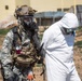 Two individuals are shown in full protective gear in an outdoor setting near a building. One person wears military digital camouflage uniform with tactical equipment, a combat helmet, and a full-face gas mask. The other person wears a white hazmat/Tyvek suit with a respirator mask, and appears to be being escorted or guided by the first individual.