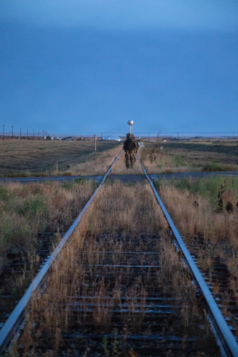 A single figure in military camouflage gear walks along railroad tracks stretching into the distance at dusk or dawn. The flat, arid landscape is flanked by dry grassland, with a small town or installation visible on the horizon, including a water tower. The low-light blue-hour conditions and receding perspective create a stark, solitary composition.